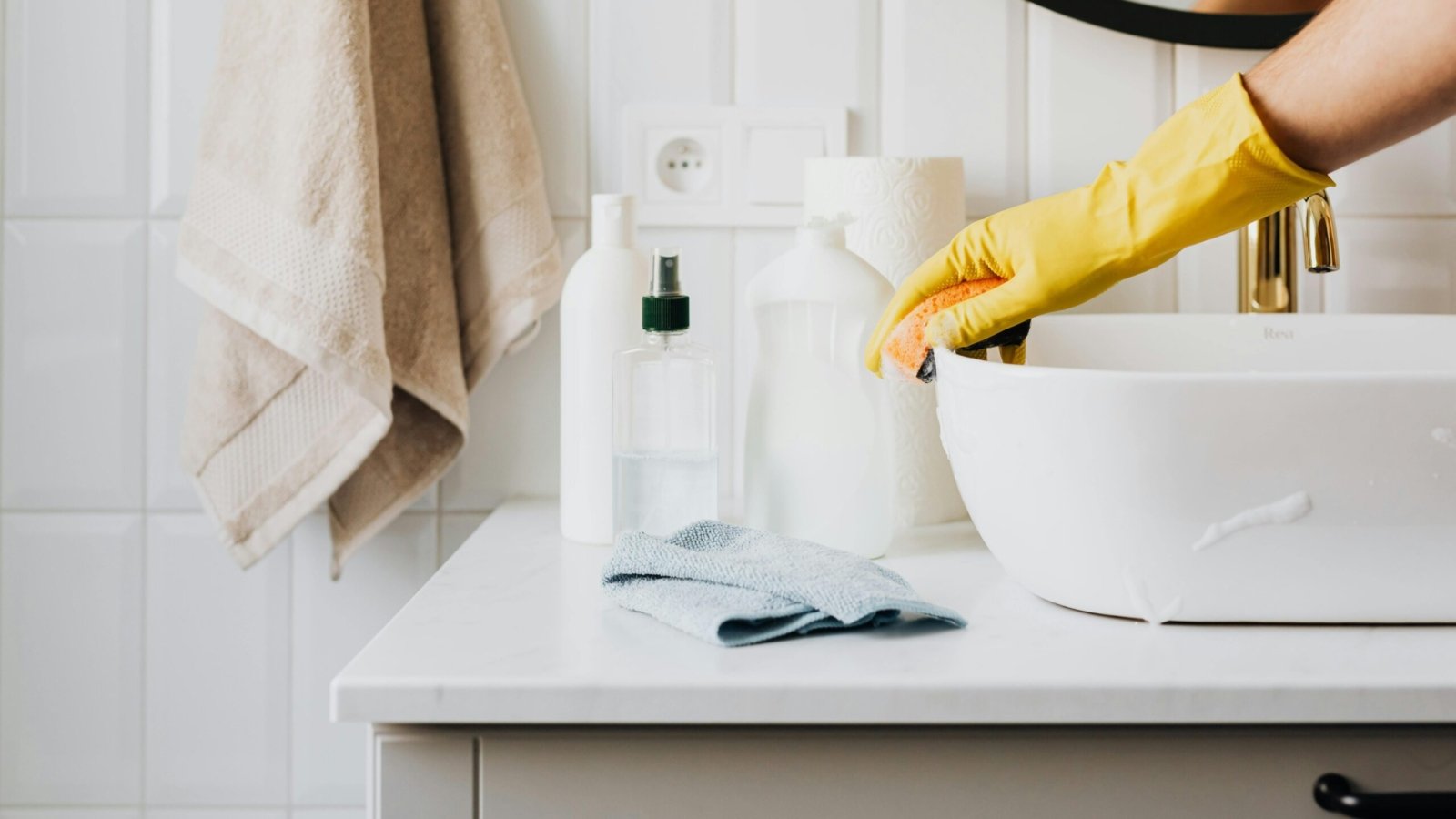 Person cleaning a bathroom sink with yellow gloves and cleaning products on a countertop.
