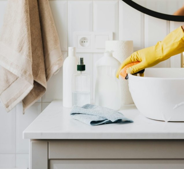 Person cleaning a bathroom sink with yellow gloves and cleaning products on a countertop.
